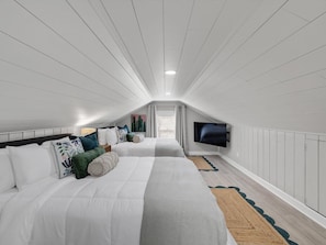 A bedroom with a sloped ceiling, a bed with multiple pillows, a television mounted on the wall, and a window with curtains.