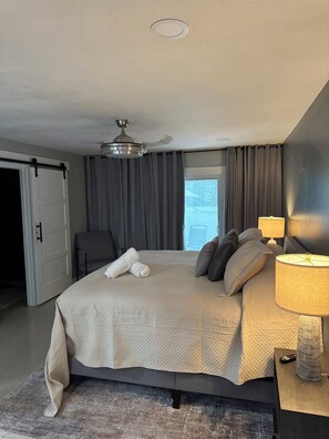 A hotel room with a bed, a desk with a lamp, a chair, a ceiling fan, and a view of the outside.