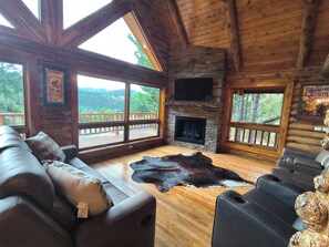 Ein Blockhaus-Interieur mit Kamin aus Stein, Ledersofa und großem Fenster mit Blick auf einen bewaldeten Bereich.