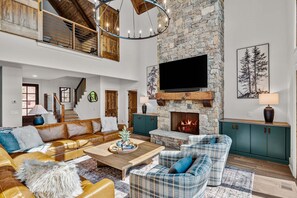 A spacious living room with a stone fireplace, a large flat-screen TV, and a high ceiling with exposed beams. - Ridge & Repeat Lodge at Eagles Nest - hot tub, in+out fireplace, firepit, views (Banner Elk)