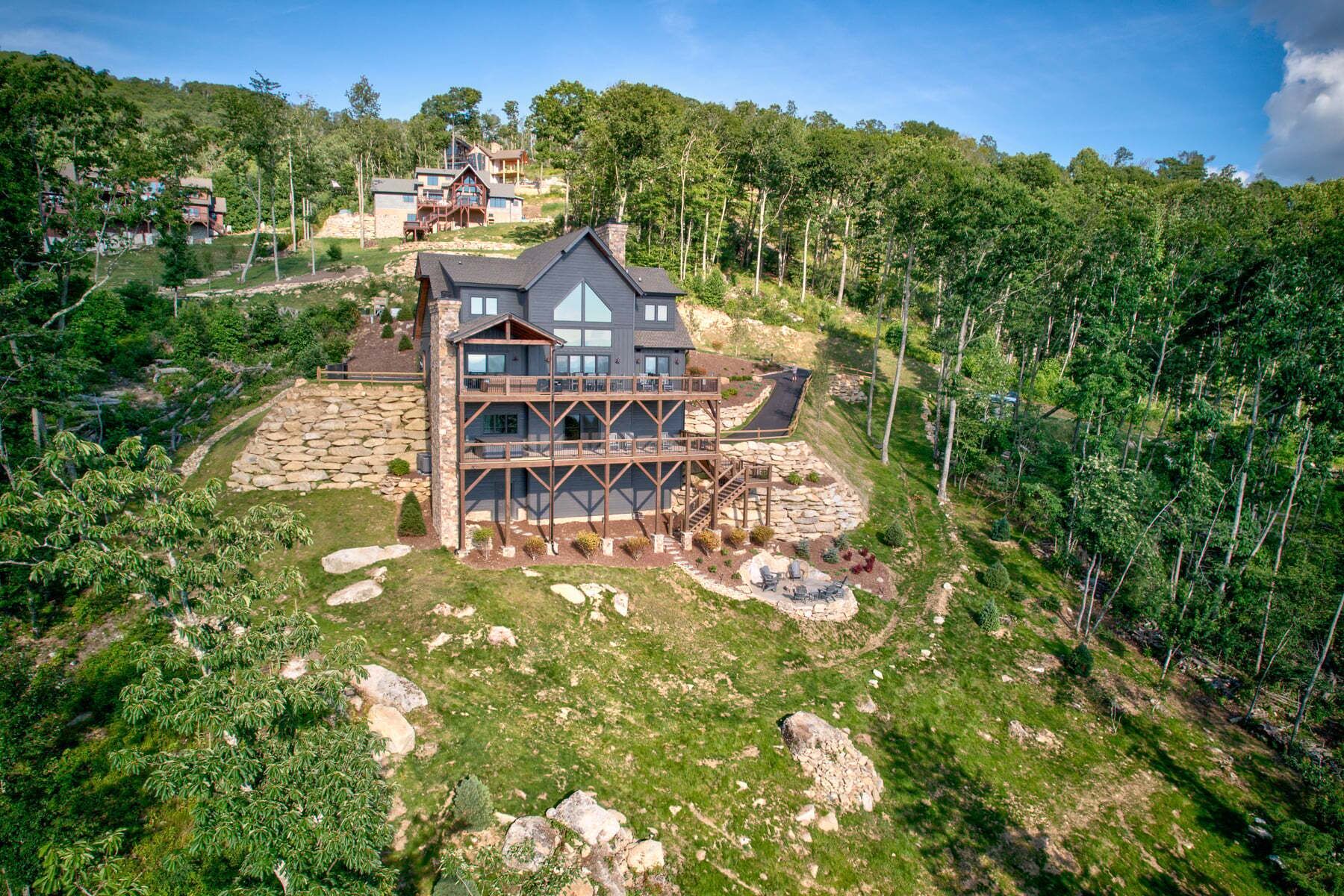 A multi-story house with a stone foundation and wooden deck, surrounded by a forested hillside.