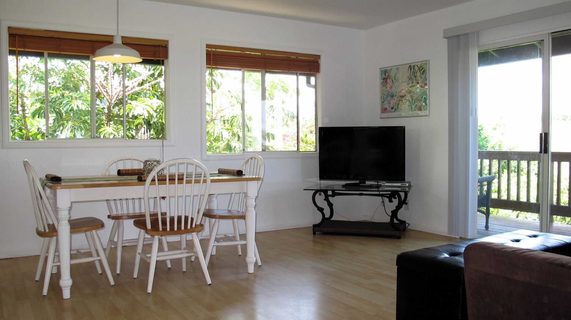 A dining area with a table and chairs, a television on a stand, and large windows with blinds.