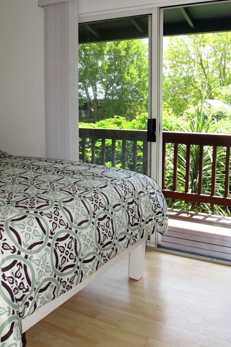 A bedroom with a bed, a sliding glass door leading to a balcony with a wooden railing, and a view of greenery outside.