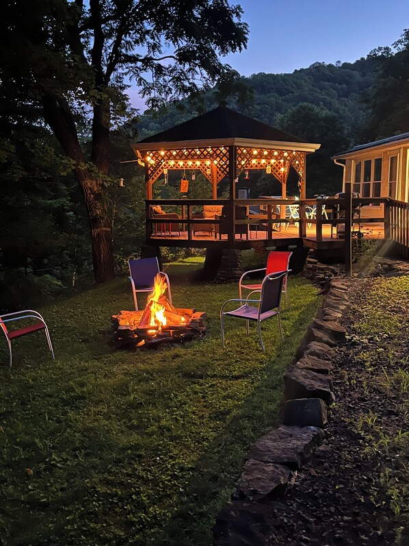 A gazebo with a fire pit and seating, surrounded by a stone border and trees.