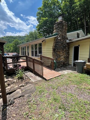 A two-story building with a stone chimney, a covered porch, and a view of a forested hillside. - Adorable 2-bedroom lake house with Lake Access near Confluence (Confluence)