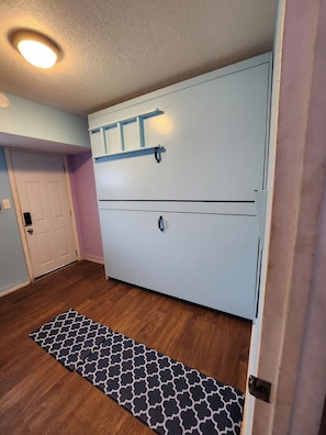 A room with a large white wardrobe, a wooden floor, a patterned rug, and a door with a light switch. - Central Maui Studio • Wall Bunks + Kitchenette (Wailuku)