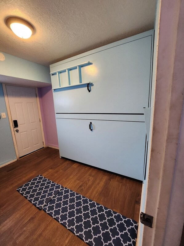 A room with a large white wardrobe, a wooden floor, a patterned rug, and a door with a light switch.