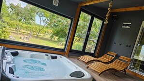 Ett spa-rum med ett bubbelbad och träloungestolar, stora fönster med utsikt över grönskan.