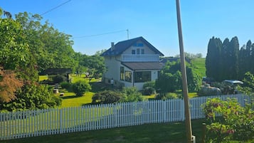 A two-story house with a balcony, surrounded by a white picket fence, in a sunny rural setting.