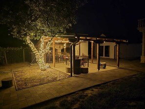 A covered outdoor seating area with a fire pit, surrounded by a wooden pergola and string lights.