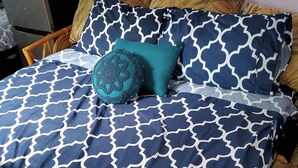 Ein Bett mit gemustertem Überwurf und Kissen, ein hölzernes Kopfteil und eine Mikrowelle auf einem Tisch links.