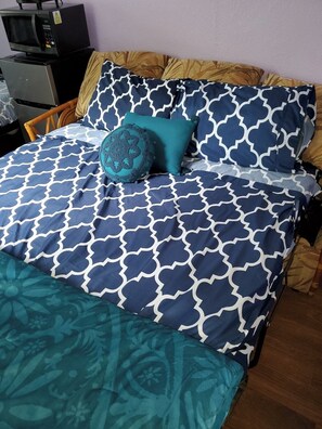 A bed with a patterned bedspread and pillows, a wooden headboard, and a microwave on a table to the left. - Downtown Wailuku Studio • Queen Bed + AC (Wailuku)