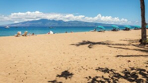 Ein Strand mit blauem Himmel, einem Gebirgszug im Hintergrund und mehreren Sonnenschirmen und Liegestühlen.