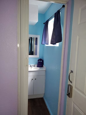 A narrow bathroom with a white sink, a mirror, and a window with a curtain. - Downtown Wailuku Studio • Queen Bed + AC (Wailuku)