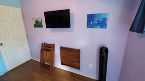 Una habitación con televisión, dos cuadros, una silla de madera, una cama y un ventilador de pie.