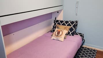 A single bed with a purple bedspread and two patterned pillows, a white headboard, and a patterned rug on a wooden floor.