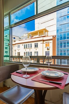 Ein Esstisch mit zwei Weingläsern und Tellern, gedeckt mit roten Platzsets, in einem Raum mit Blick auf ein Gebäude durch ein Fenster.