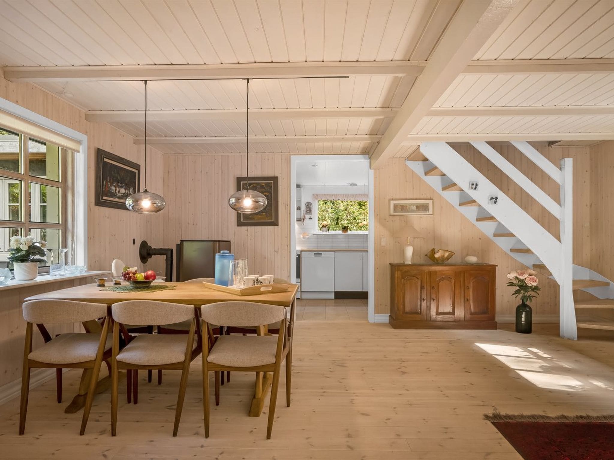 Un coin repas avec une table et des chaises en bois, une cuisine à l’arrière-plan et un escalier menant à un niveau supérieur.