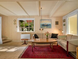 Ein gemütliches Wohnzimmer mit Holzboden, einem roten Teppich mit Muster, einem Holztisch, einer Couch und einem Fenster mit Blick ins Grüne.