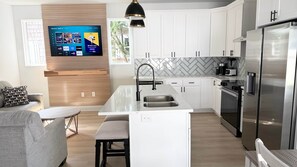 Eine moderne Küche mit weißen Schränken, einer zentralen Kochinsel, Edelstahlgeräten und einem an der Wand befestigten Flachbildfernseher.