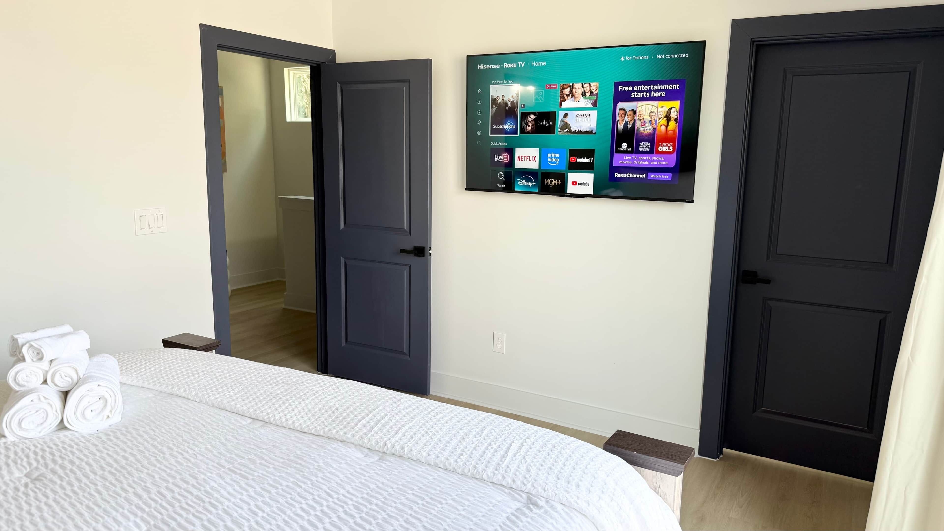 A hotel room with a bed, a wall-mounted TV displaying streaming services, and a door leading to another room.