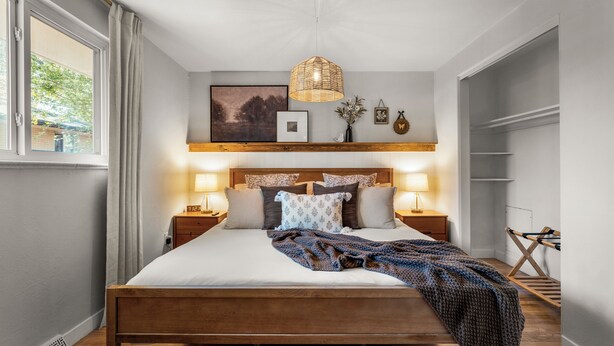 A neatly arranged bedroom with a wooden bed, bedside lamps, a wall-mounted shelf with decorative items, and a window with curtains. - Dog-Friendly Arvada Home with Fire Pit, 18 Min to Red Rocks & Near Denver (Arvada)