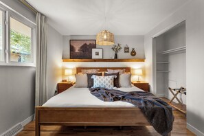 A neatly arranged bedroom with a wooden bed, bedside lamps, a wall-mounted shelf with decorative items, and a window with curtains. - Dog-Friendly Arvada Home with Fire Pit, 18 Min to Red Rocks, King Bed, & Games (Arvada)