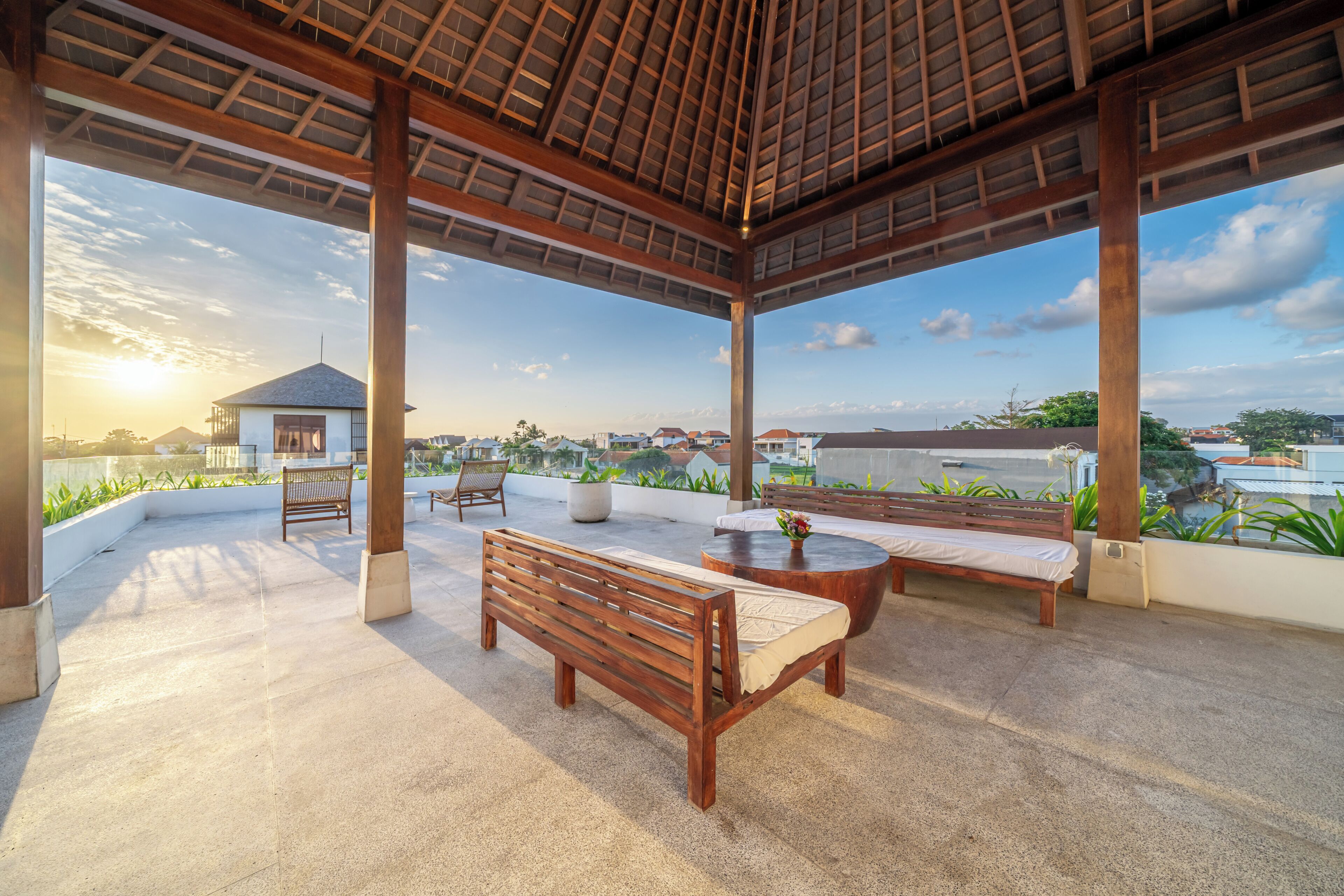 A covered outdoor seating area with benches and a table, overlooking a residential area with houses and a body of water.