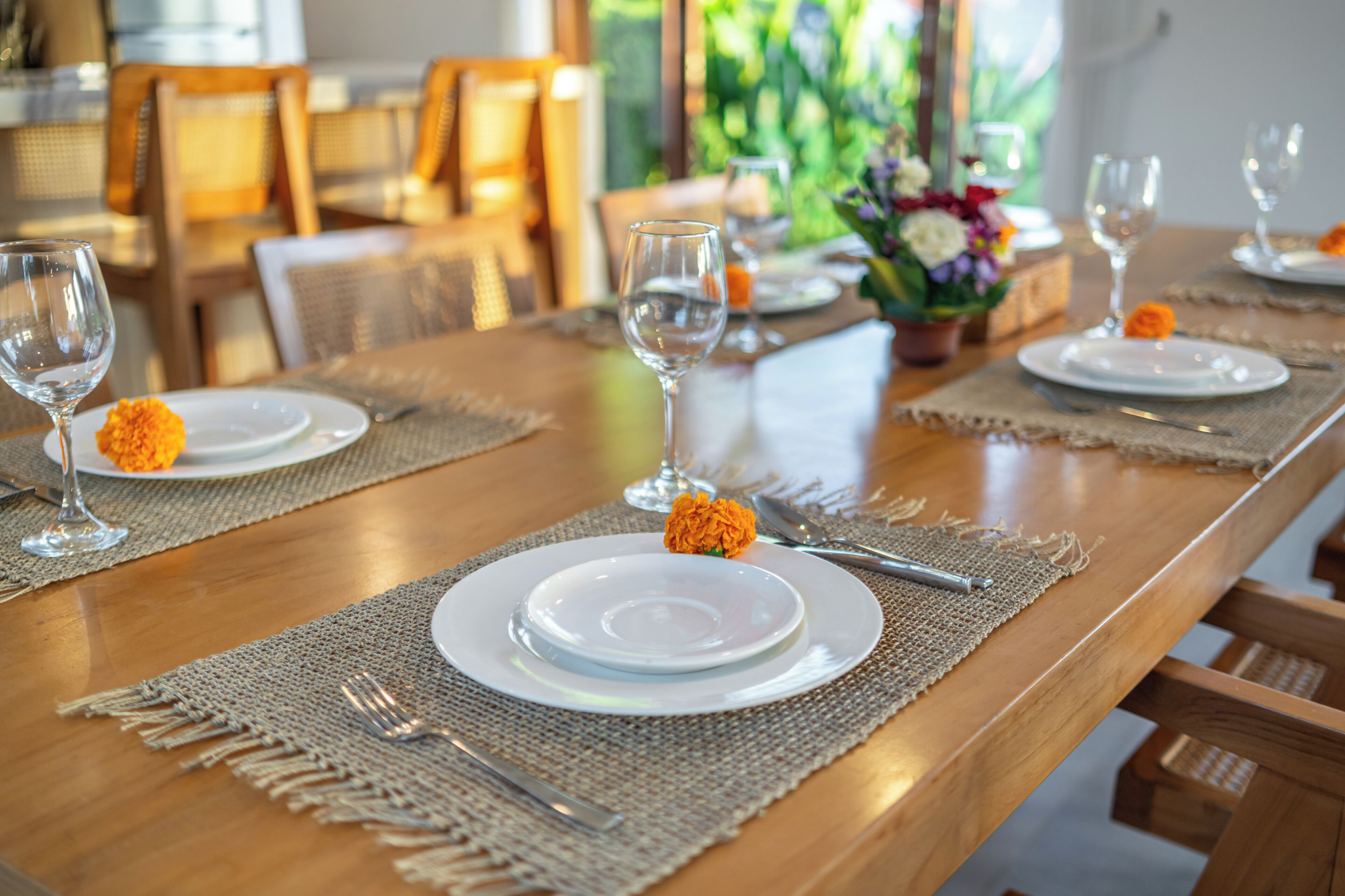 Une table à manger dressée avec de l’assiette blanche, des couverts et des verres à vin, agrémentée d’un set de table en fibres tressées et d’une seule fleur orange.