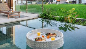 Une table à manger avec un panier en osier contenant de la nourriture et des boissons, une piscine et une vue sur un quartier résidentiel avec des maisons et de la verdure.