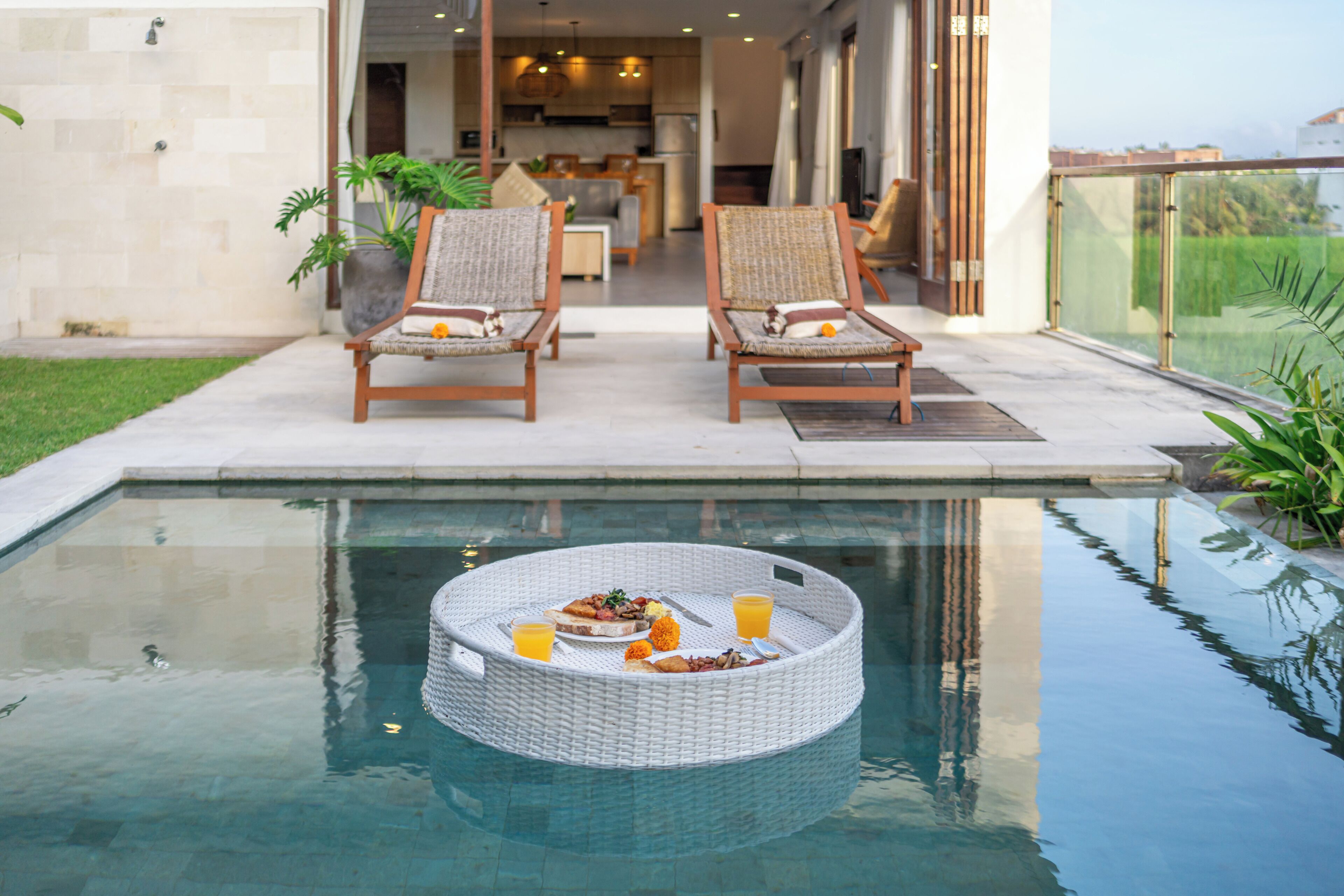 Uma área de refeições à beira da piscina, com duas cadeiras de vime, uma mesa redonda com comida e bebidas, e vista desobstruída para a piscina.
