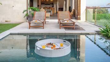 Uma área de refeições à beira da piscina, com duas cadeiras de vime, uma mesa redonda com comida e bebidas, e vista desobstruída para a piscina.