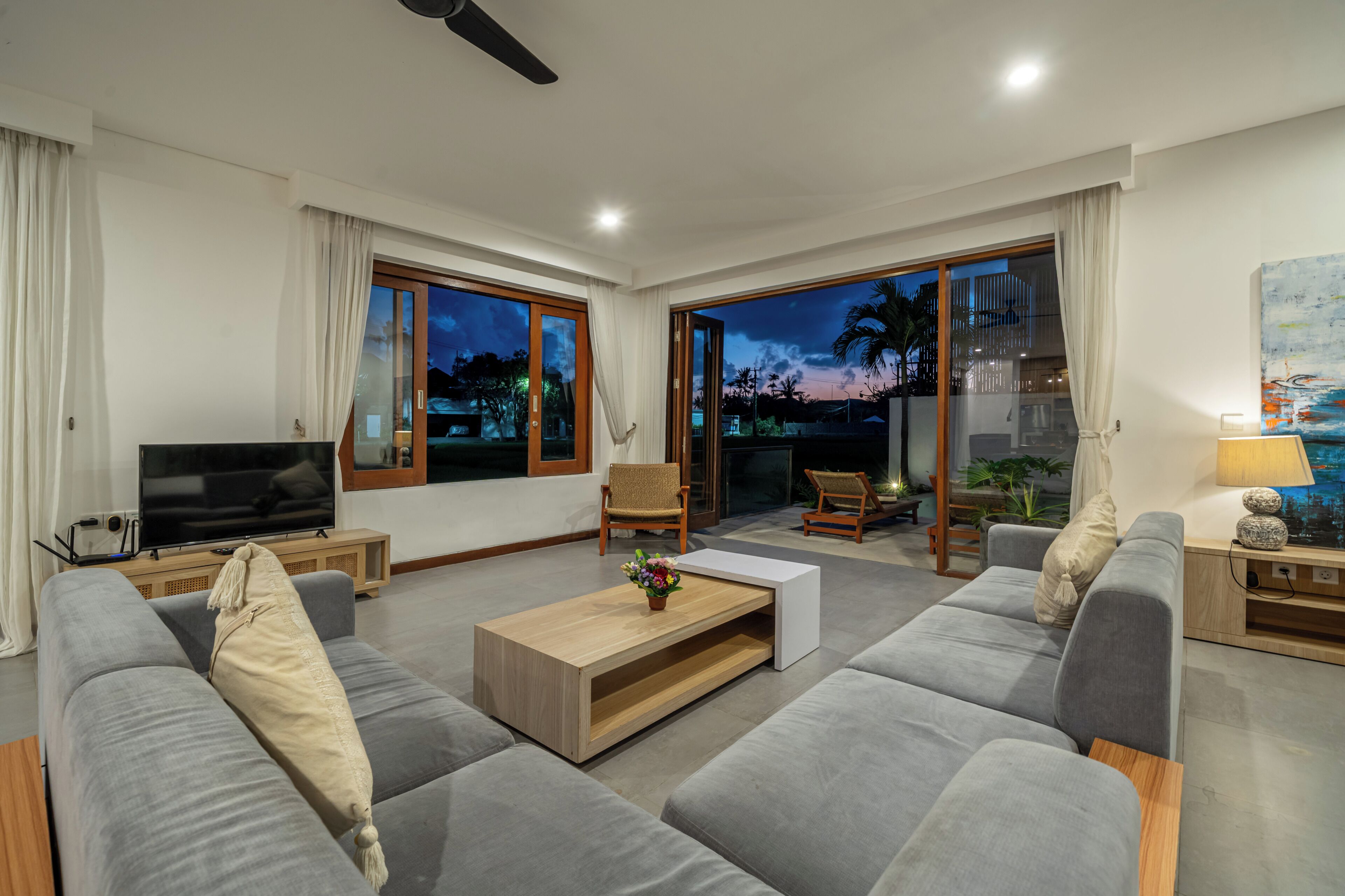A modern living room with a sofa, a coffee table, and a television. There is a sliding glass door leading to an outdoor area with a view of the sky and palm trees.