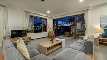 A modern living room with a sofa, a coffee table, and a television. There is a sliding glass door leading to an outdoor area with a view of the sky and palm trees.