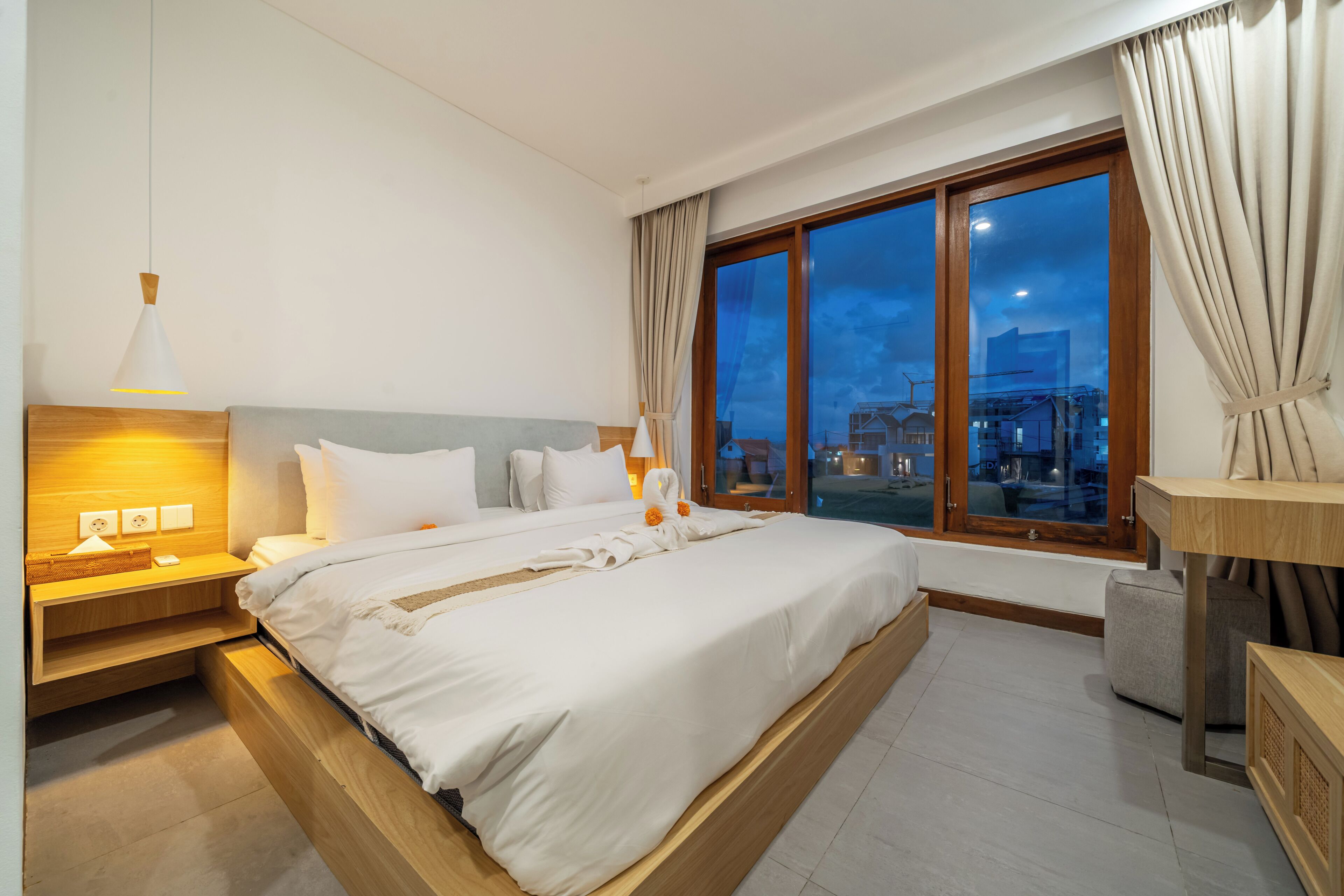 A modern hotel room with a large bed, bedside tables, a nightstand, a lamp, and a view of the city at dusk.