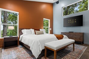 Un dormitorio moderno con una cama grande, un banco, un televisor y vistas a árboles a través de las ventanas.