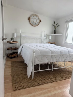 Ein Schlafzimmer mit einem weißen Metallbettgestell, einem Nachttisch aus Holz mit einer Lampe, einer kleinen Pflanze und einem wandmontierten Regal.