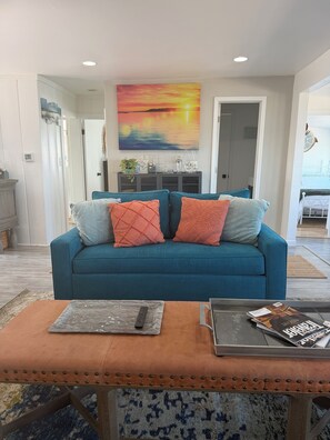 A living room with a teal sofa, orange cushions, a coffee table with magazines, and a painting of a sunset.