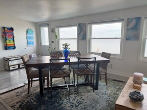 A dining area with a wooden table and chairs, a vase with a plant, and a view of the ocean through large windows. - Lake Huron Cottage - Walkout Beachfront - Sleeps 8 (Kawkawlin)