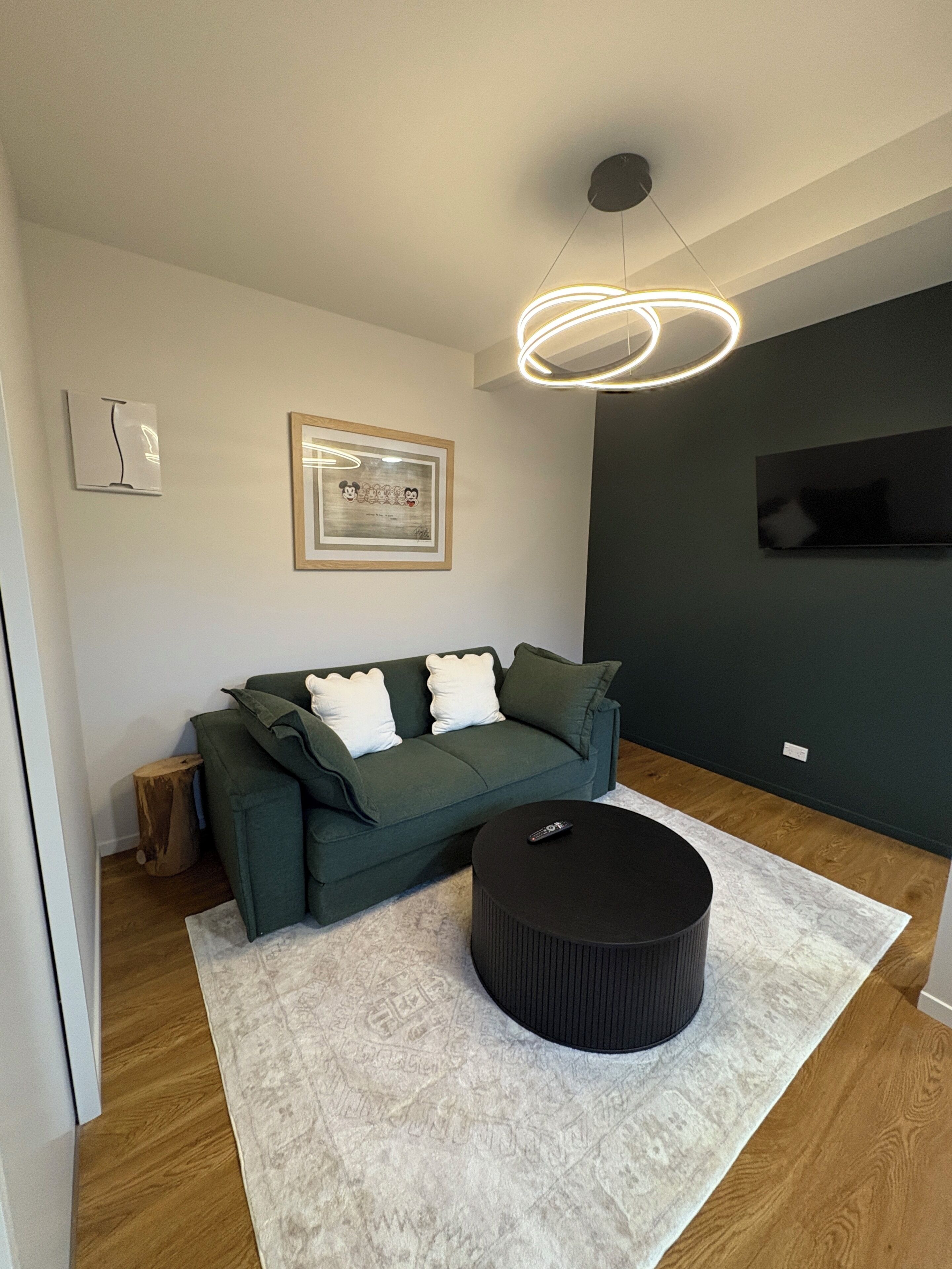 A modern living room with a green sofa, a round ottoman, a framed picture on the wall, and a ceiling light fixture.