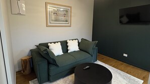 Ein modernes Wohnzimmer mit einem grünen Sofa, einem runden Hocker, einem gerahmten Bild an der Wand und einer Deckenleuchte.