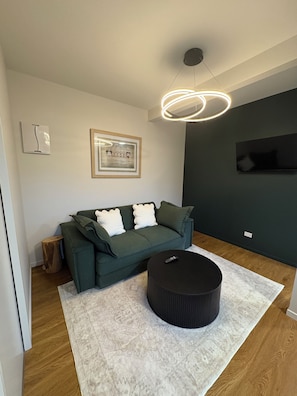 A modern living room with a green sofa, a round ottoman, a framed picture on the wall, and a ceiling light fixture.
