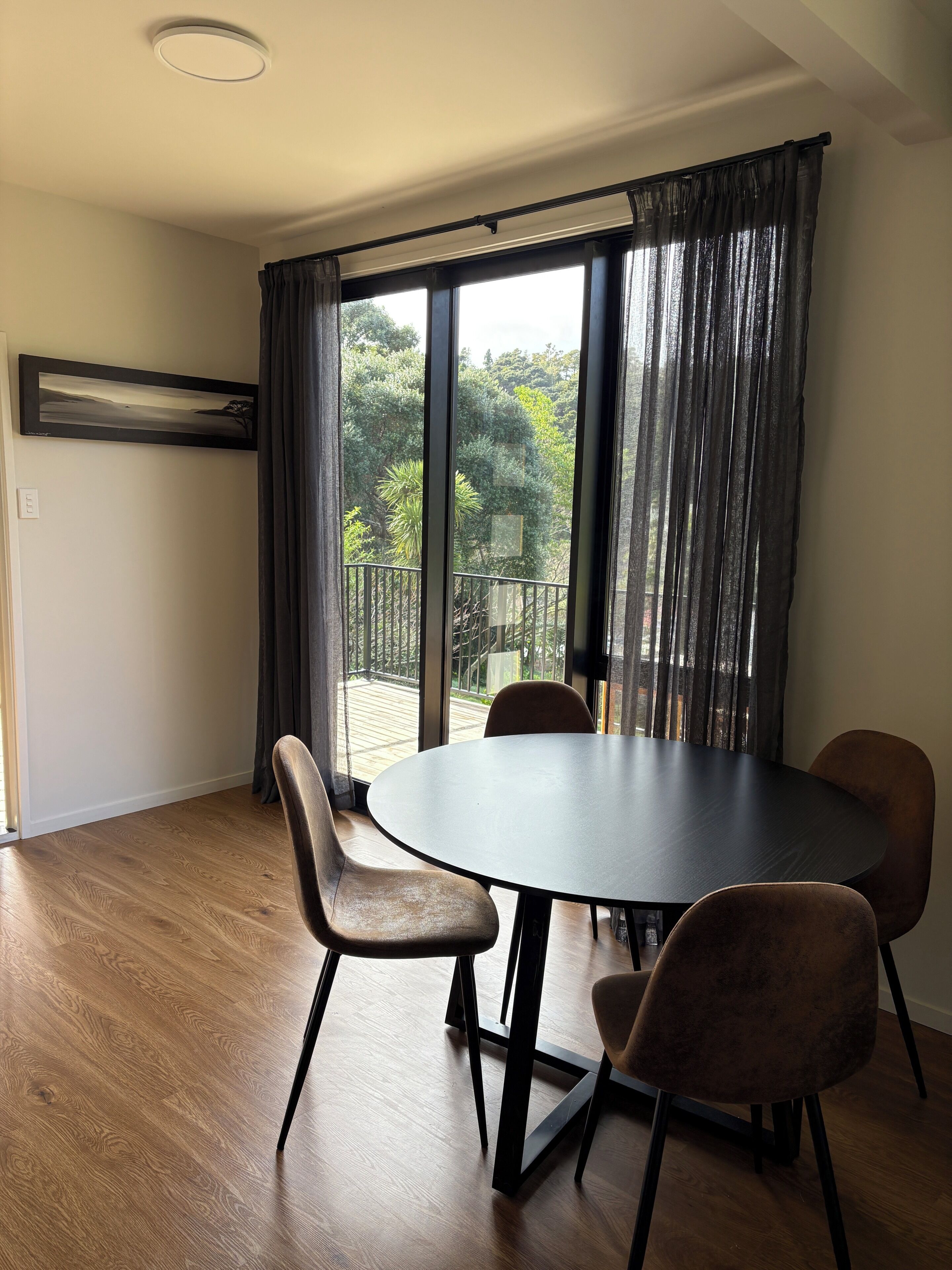 A dining area with a round table and four chairs, large windows with curtains, and a view of greenery outside.
