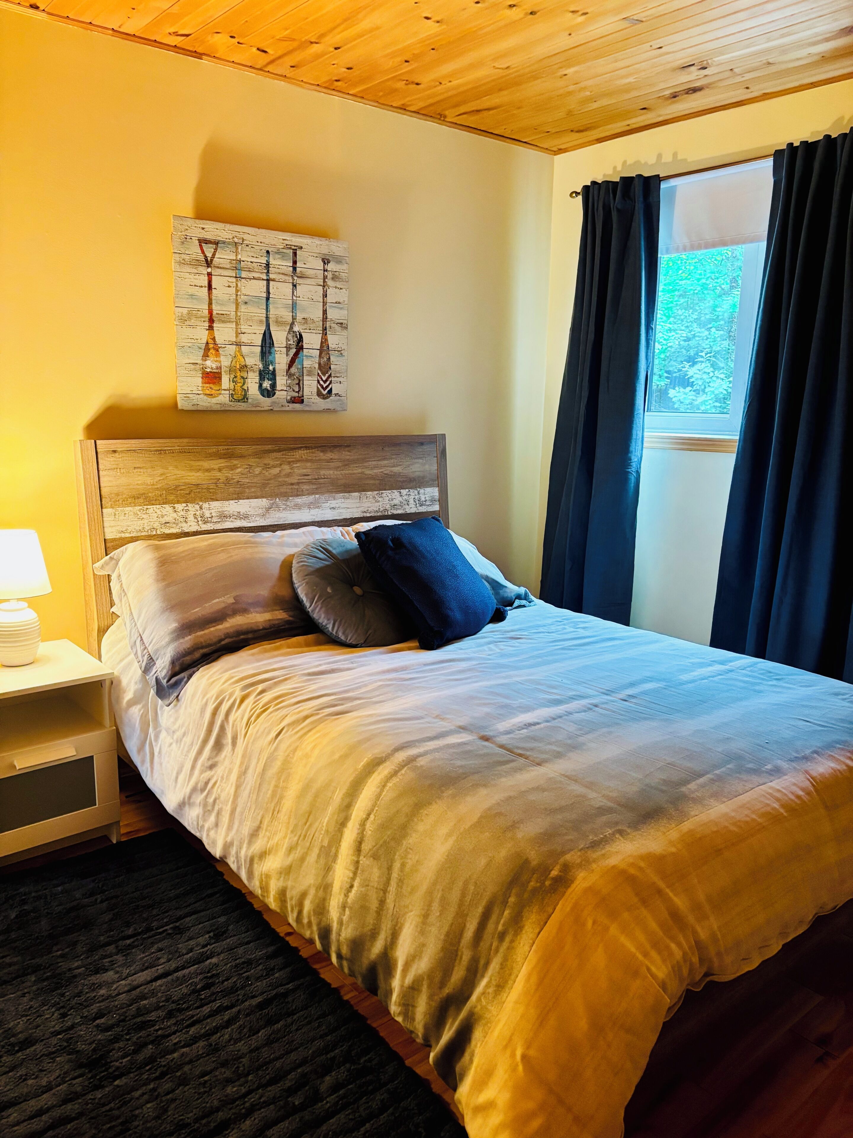 Een slaapkamer met een houten hoofdbord, een bed met een beige en grijze dekbedovertrek, een nachtkastje met een lamp, en een raam met donkere gordijnen.