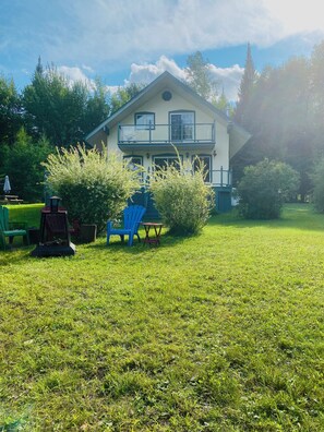A two-story house with a balcony, surrounded by a grassy lawn and trees. - The Secret of the Lake (Rivière-Rouge)