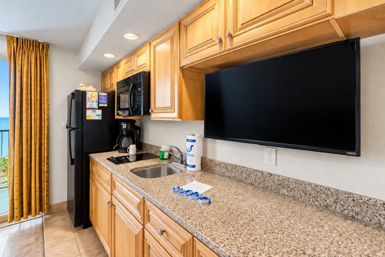 A kitchen with wooden cabinets, a black refrigerator, a microwave, a coffee maker, and a flat-screen TV.