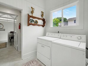 A laundry room with a large washer and dryer, a wooden shelf with decorative items, and a window with a view of a house and trees. - Coastal Grand 7 bed, 4 bath + hot tub & fire pit (Ludington)