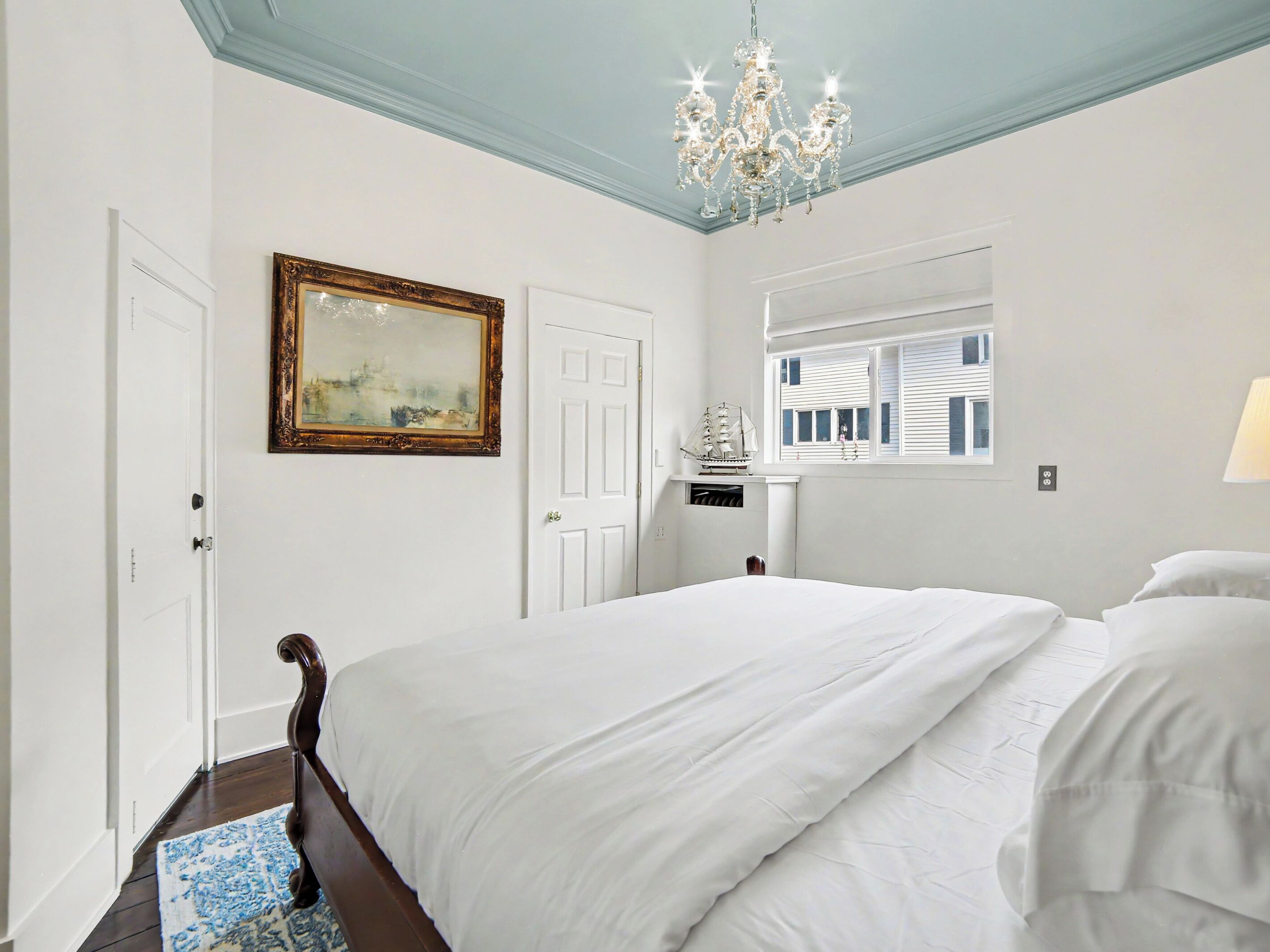 A bedroom with a large bed, a chandelier, a window with shutters, and a painting on the wall.