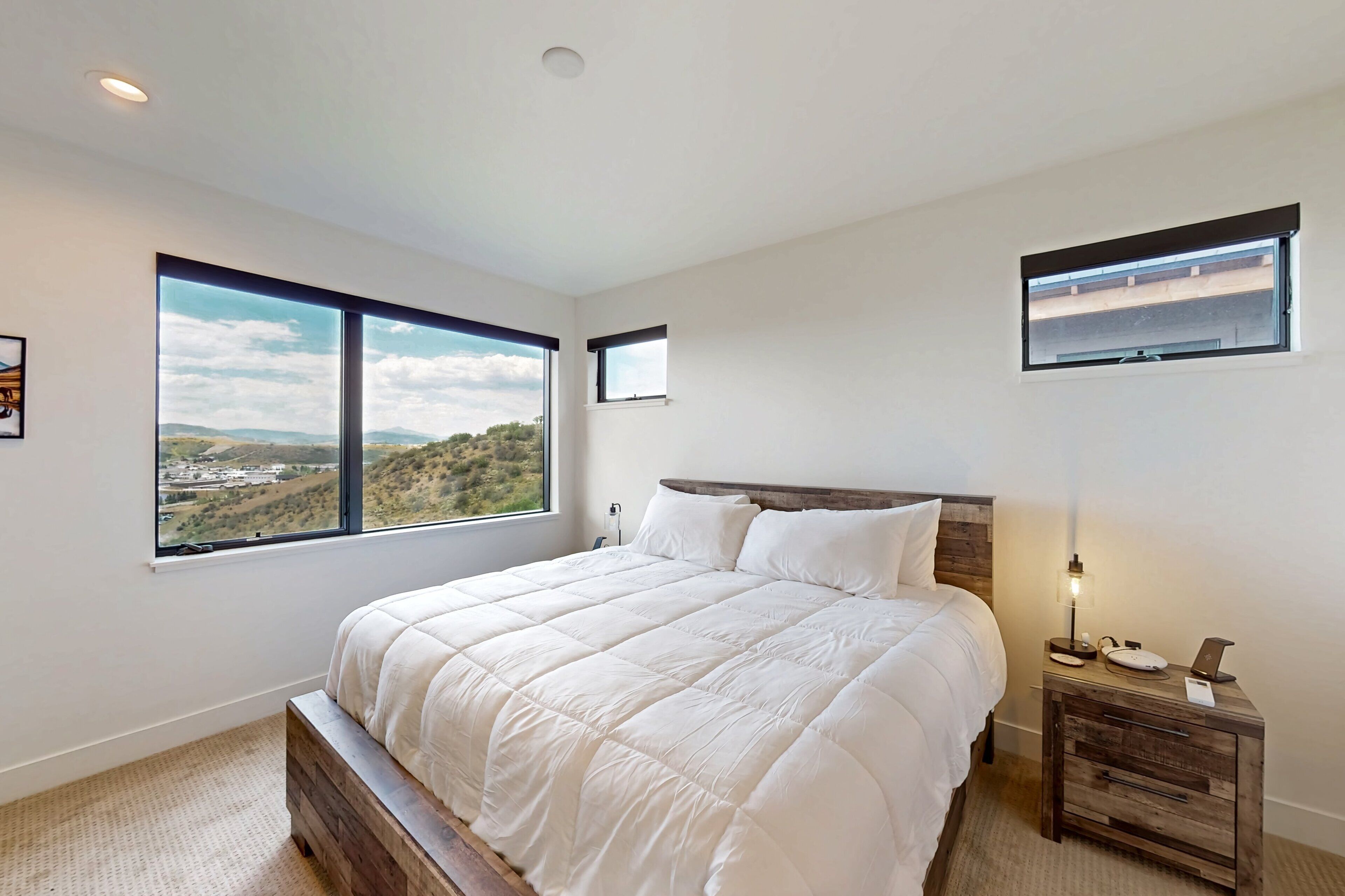 A bedroom with a large bed, a nightstand, a lamp, and a view of a landscape through a window.