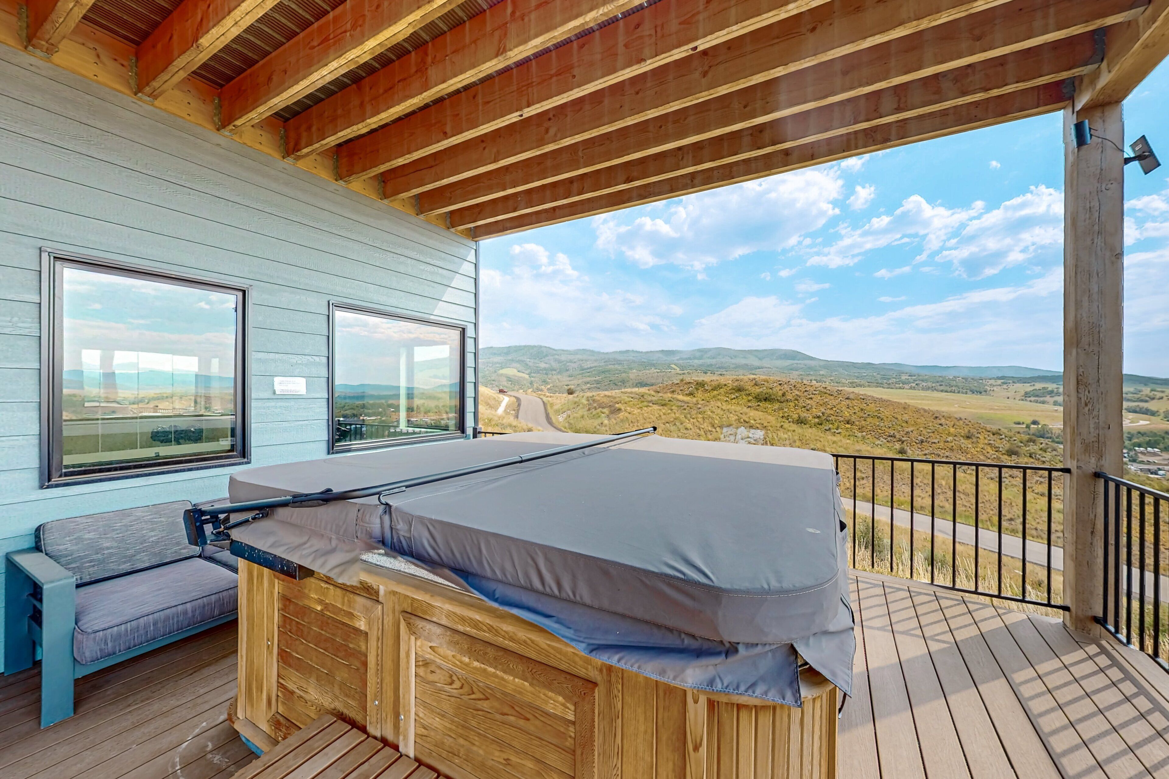 A wooden deck with a bed and a bench, overlooking a landscape with hills and a road.